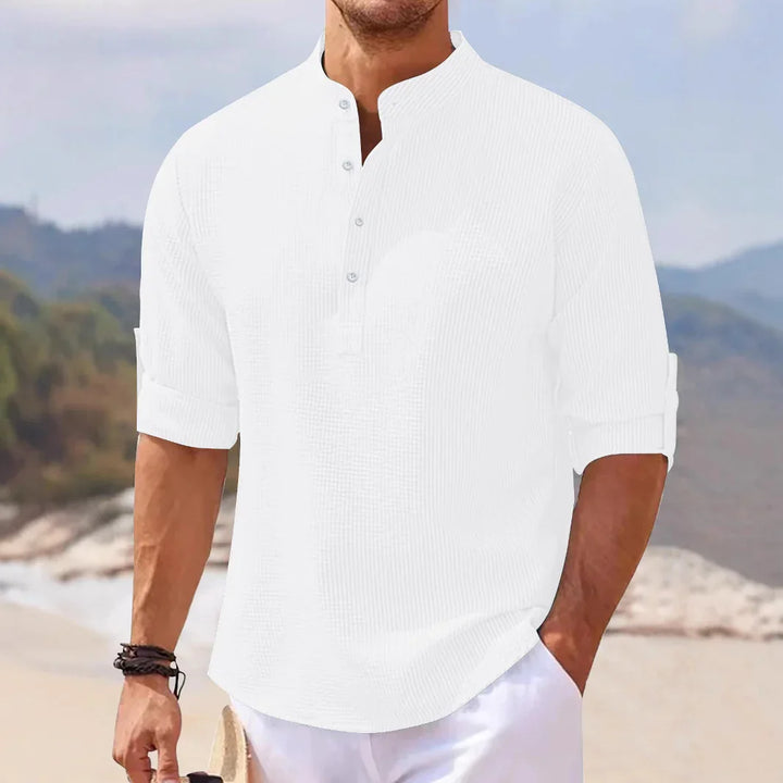 Man wearing white casual linen shirt with rolled sleeves standing on a beach with hills in the background
