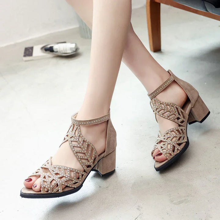 Beige suede block heel sandals with ankle strap and studded geometric cutout design on feet