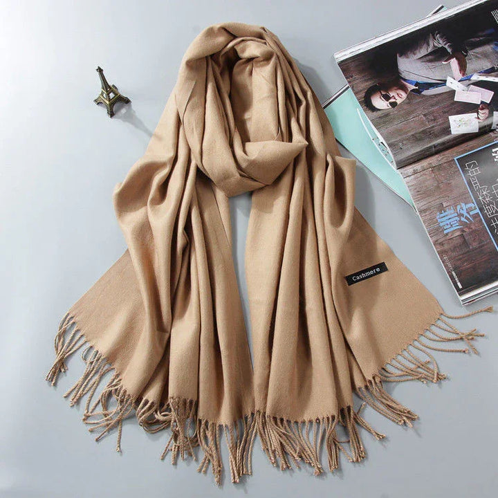 Beige cashmere scarf with fringed edges laid flat next to a mini Eiffel Tower model and opened magazine