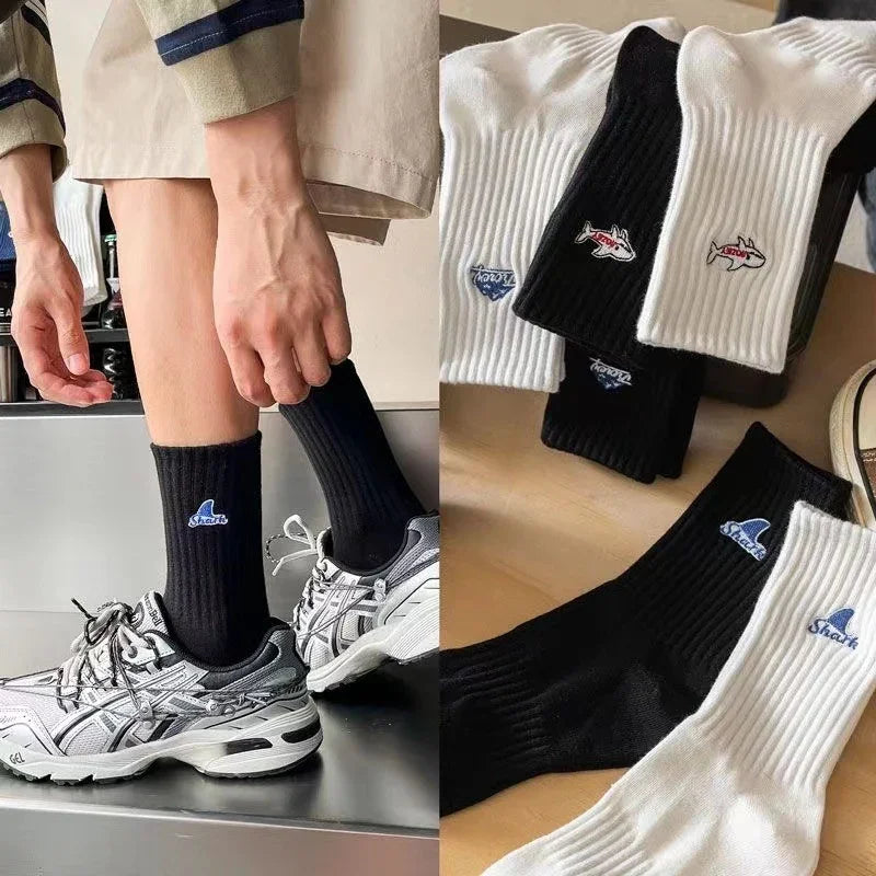 Person wearing black ribbed socks with shark logo paired with gray sneakers, plus white and black shark graphic socks on a wooden surface