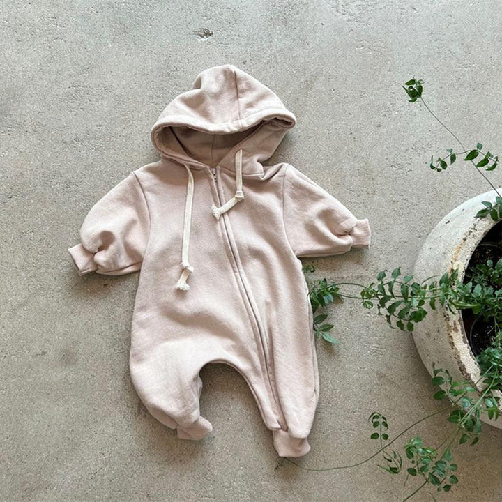 Beige baby hooded jumpsuit with front zipper and drawstring on concrete floor beside green potted plant