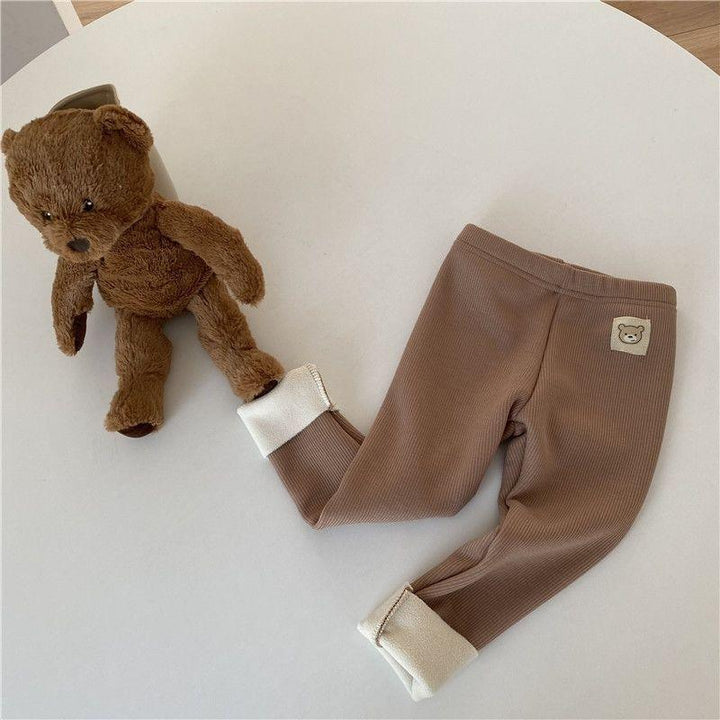 Brown ribbed baby leggings with teddy bear patch next to plush teddy bear on white surface