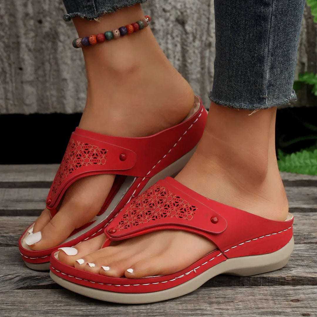 Close-up of feet wearing red open-toe sandals with decorative cutouts and beaded anklet on wooden floor