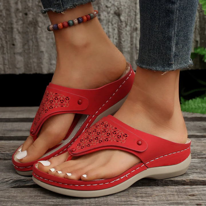 Close-up of feet wearing red open-toe sandals with decorative cutouts and beaded anklet on wooden floor