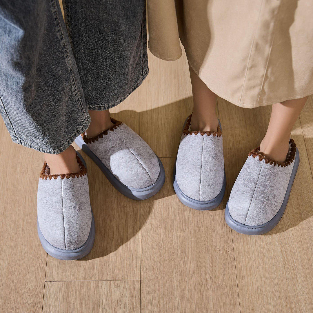 Two pairs of light gray slippers with brown trim worn by two people on wooden floor