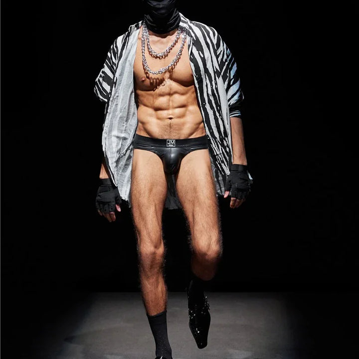Male model in black leather briefs, zebra-print open shirt, chains, gloves, and black face mask on dark runway