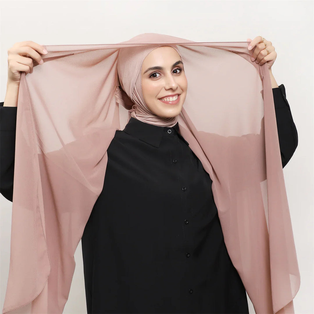 Smiling woman wearing a light pink hijab and black button-up shirt holding her scarf's ends