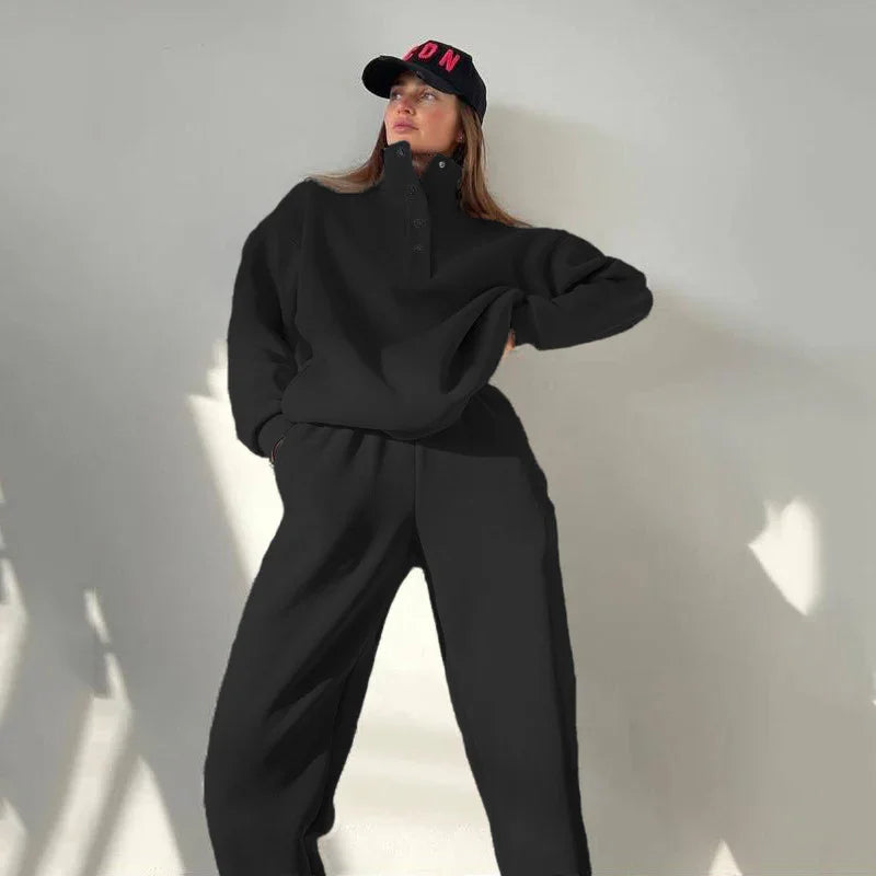 Woman in black oversized sweatshirt and pants with black cap posing against neutral wall