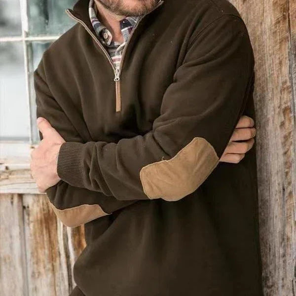 Man wearing brown quarter-zip sweater with tan elbow patches over plaid shirt, rustic wooden background