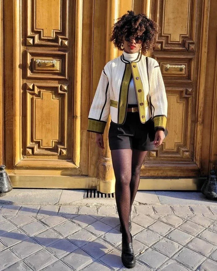 Stylish woman wearing black shorts, patterned tights, and a white jacket with yellow trim walking on cobblestone street in front of ornate wooden doors