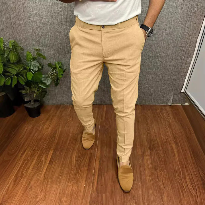 Man wearing beige slim-fit pants and brown loafers standing on wooden floor with indoor plants