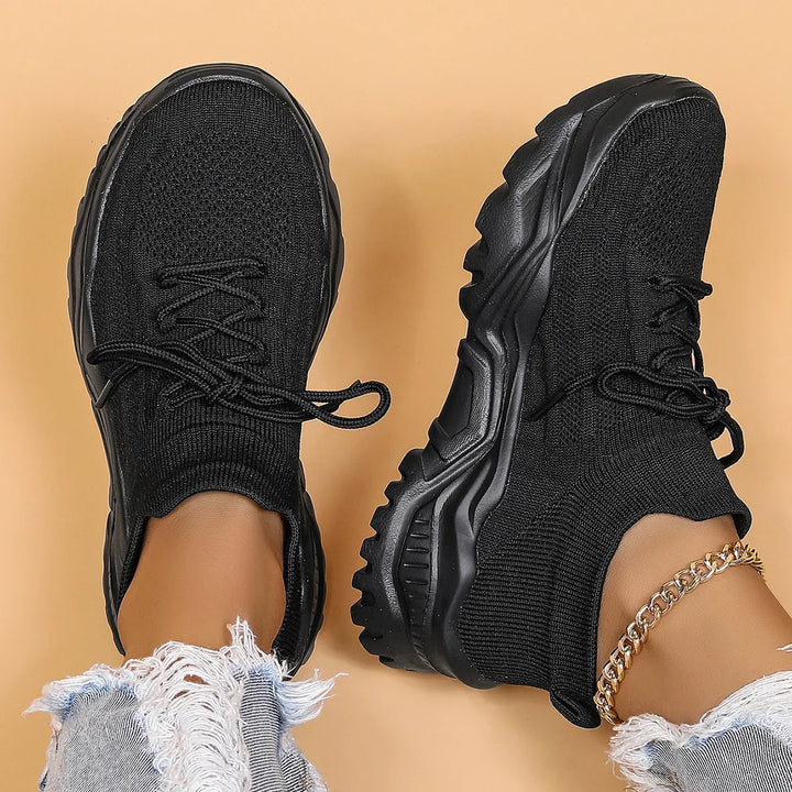 Black knit sock sneakers with rugged sole worn with ripped denim jeans and gold chain anklet on beige background