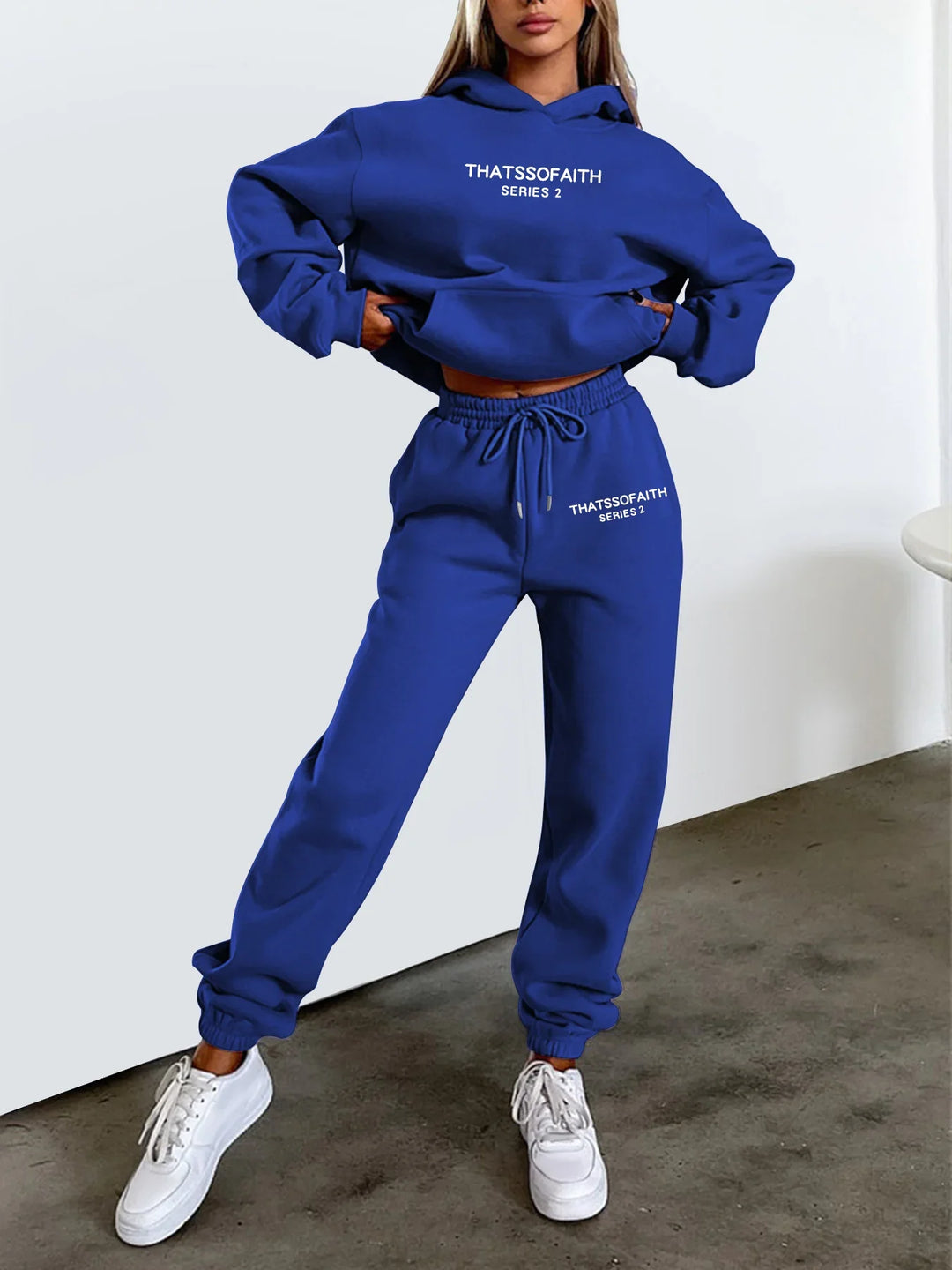 Woman wearing blue THATSSOFAITH Series 2 hoodie and jogger set with white sneakers against white background