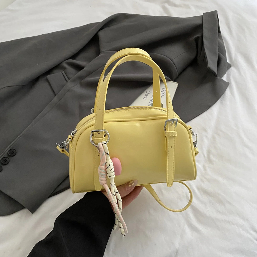 Hand holding small yellow leather handbag with silver buckles and rope charm, gray blazer on white bed