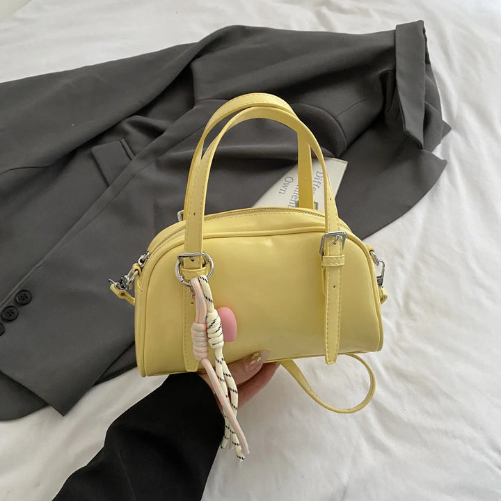 Hand holding small yellow leather handbag with silver buckles and rope charm, gray blazer on white bed