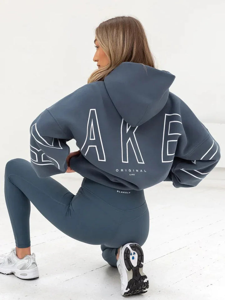 Woman wearing blue Blakely hoodie with large white lettering and matching leggings, white sneakers, indoor studio