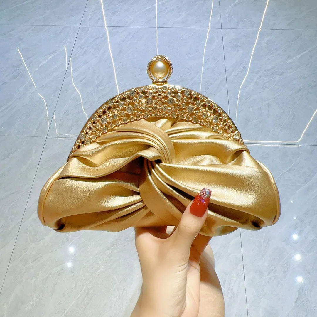 Hand holding a gold satin clutch purse with jeweled top clasp on glossy marble floor