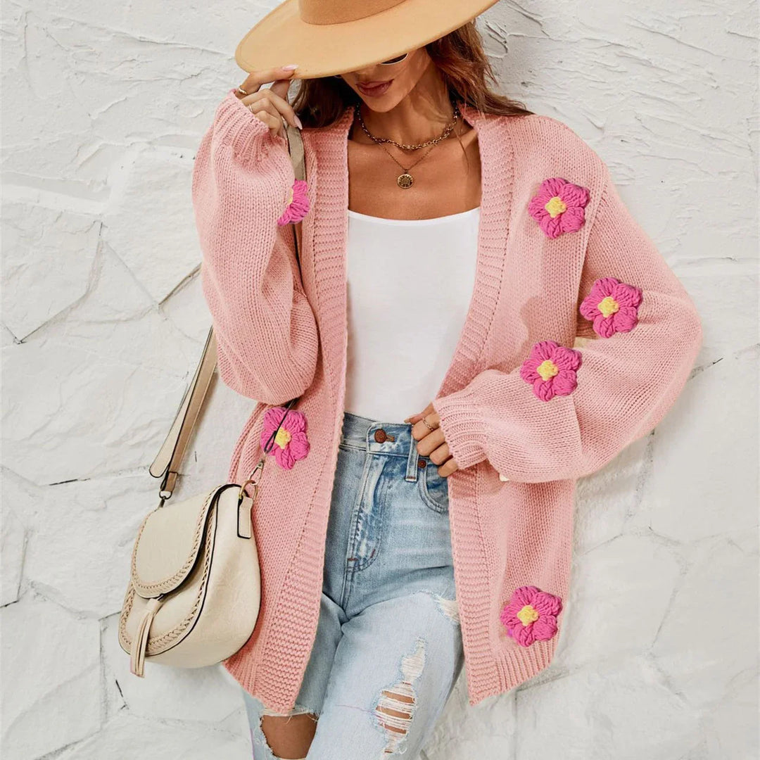 Woman wearing pink knit cardigan with floral patches, white tank top, ripped jeans, beige hat, and cream shoulder bag