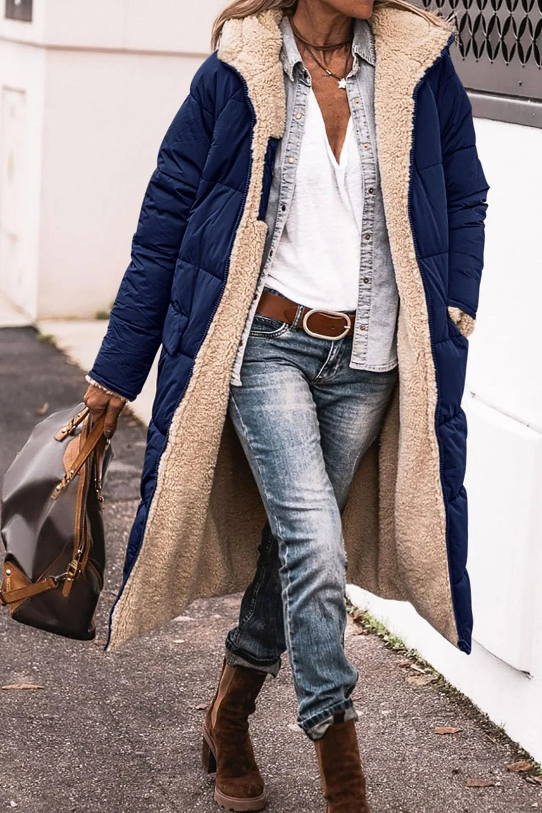 Woman wearing long navy blue sherpa-lined winter coat, denim shirt, white tee, jeans, brown boots, and carrying brown handbag