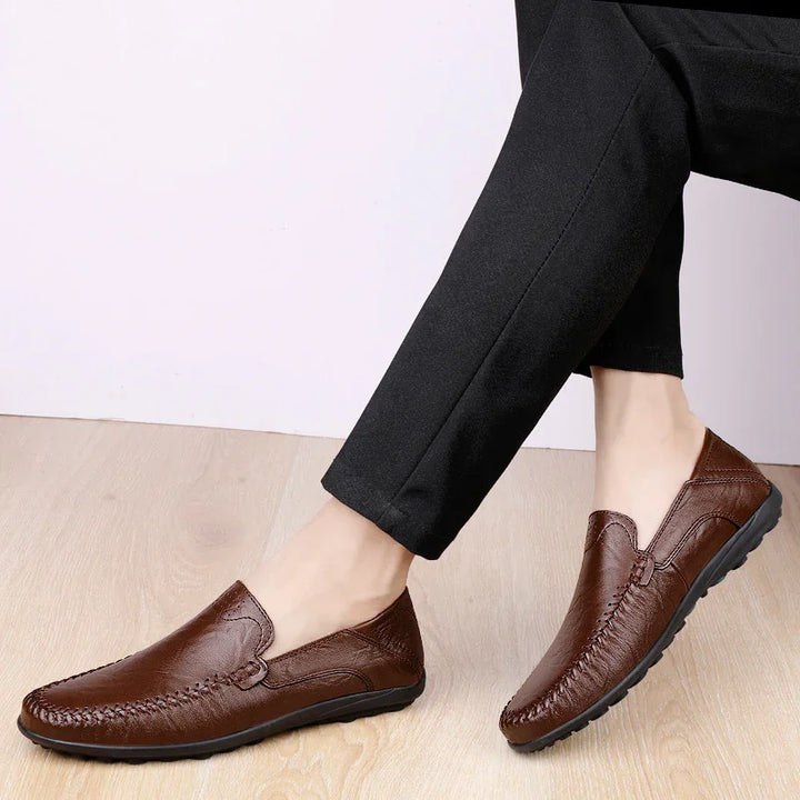 Men's brown leather slip-on casual loafers worn with black pants on light wooden floor