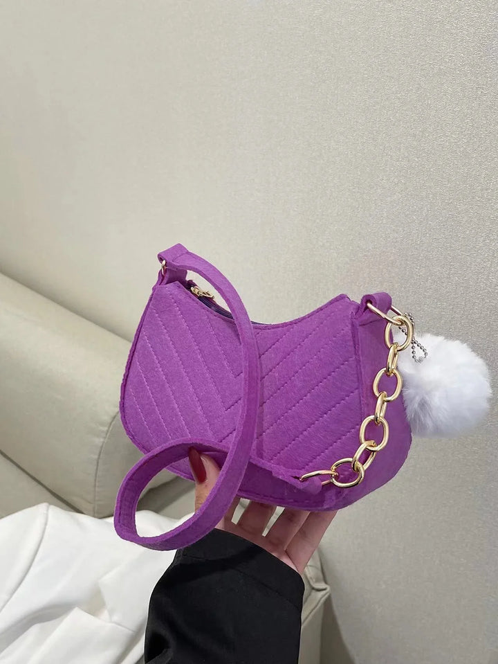 Hand holding a purple quilted small shoulder bag with gold chain and white fluffy pompom