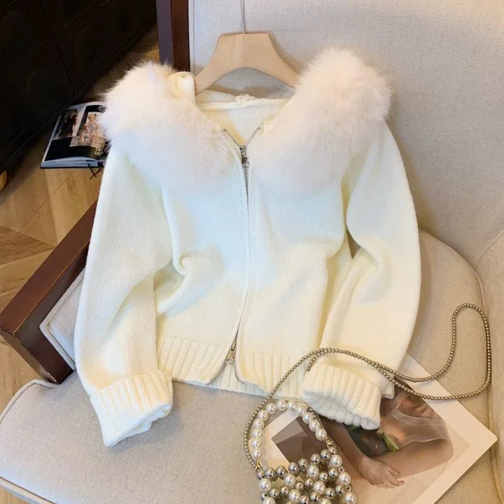 White knitted zip-up cardigan with fluffy fur collar on beige chair with pearl chain handbag