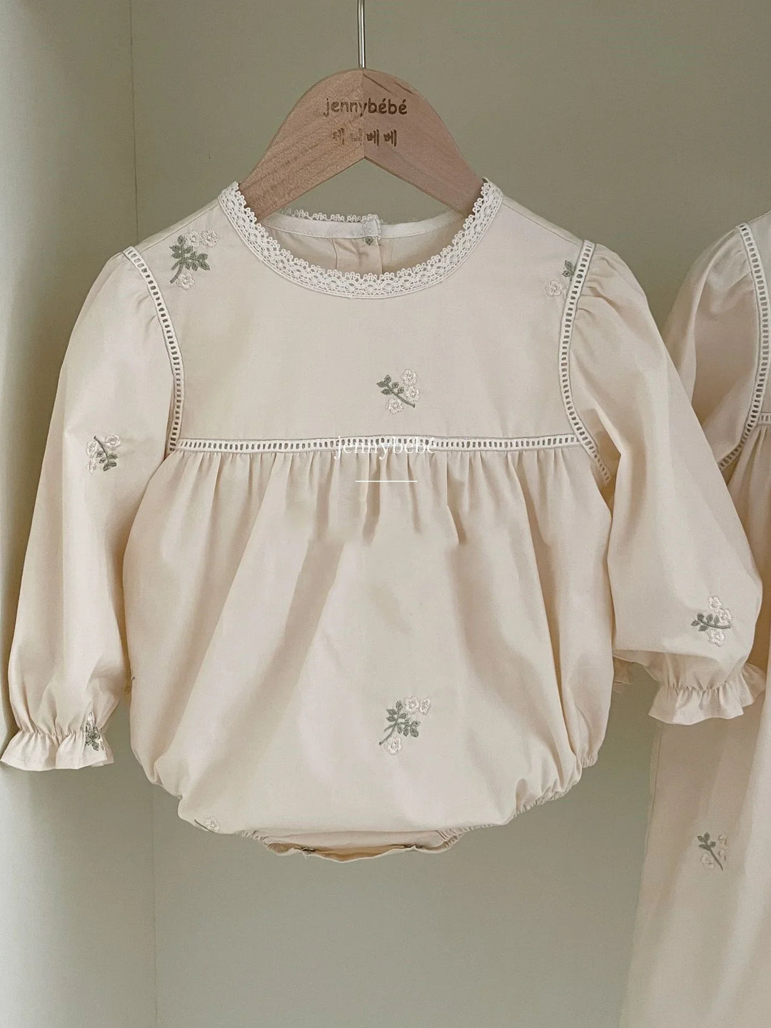 Cream baby romper with long sleeves and delicate floral embroidery, hanging on wooden hanger