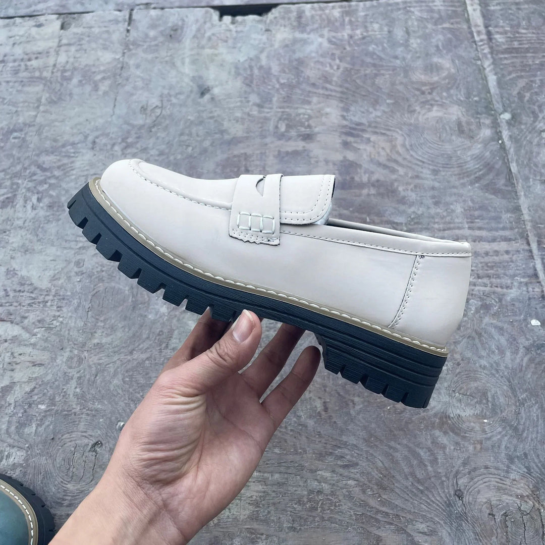 Hand holding a white leather loafer with black chunky sole on gray textured floor