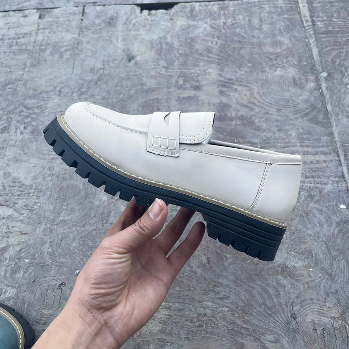 Hand holding a white leather loafer with black chunky sole on gray textured floor