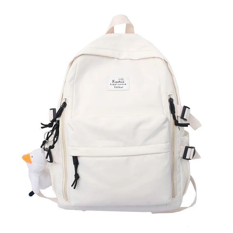White backpack with front zipper pockets, black zipper pulls, side buckles, and a small plush duck keychain