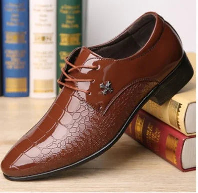 Brown glossy men's dress shoe with reptile texture and decorative metal embellishment, displayed on books