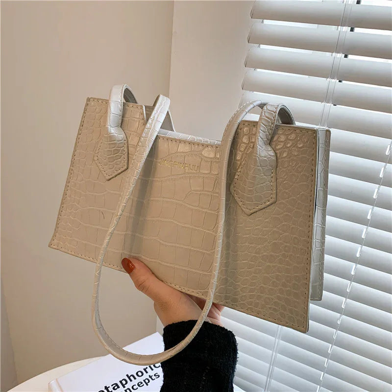 Hand holding beige crocodile texture leather handbag with thin shoulder strap near window blinds