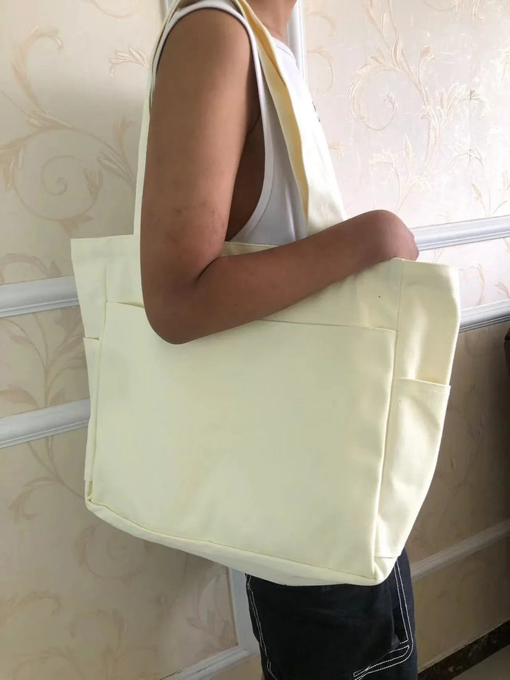 Person carrying a large plain cream-colored canvas tote bag with side pockets indoors