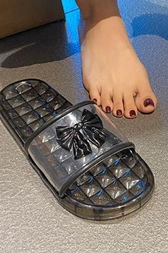 Black slide sandal with transparent strap and bow detail next to a foot with dark manicure on gray floor