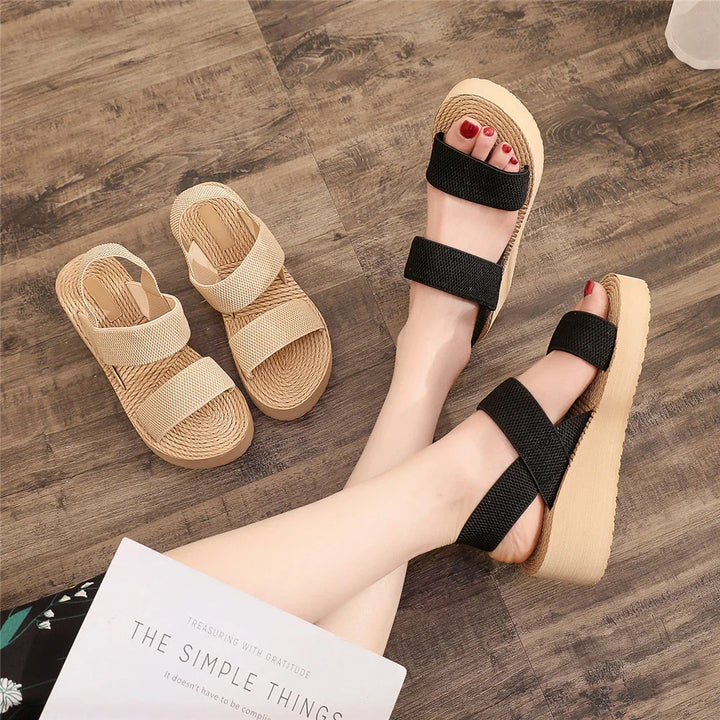 Woman wearing black double-strap platform sandals on wooden floor with beige sandals nearby