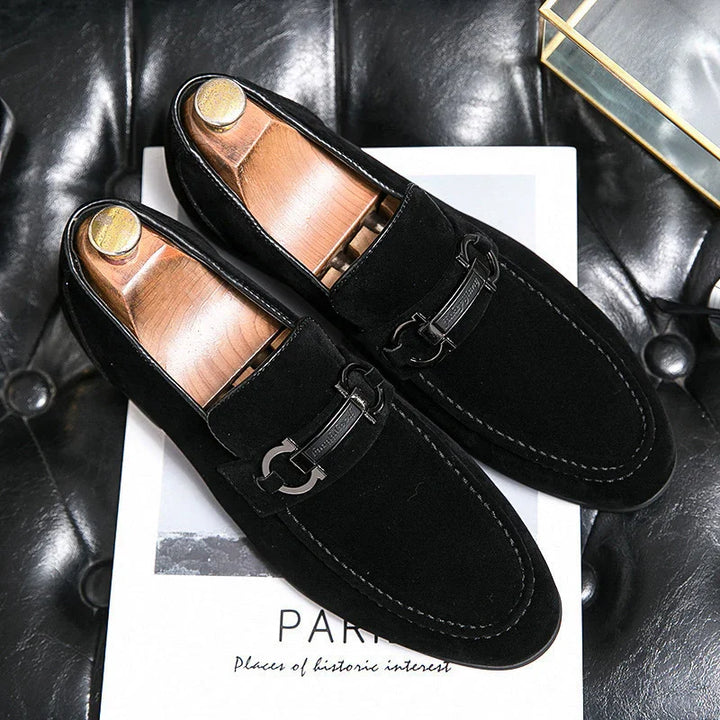 Black suede men's loafers with metal bit detail on top, displayed on a white book on a black leather surface