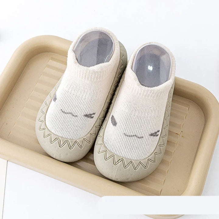 Pair of beige baby shoes with white knitted uppers and cute face design, placed on tan tray