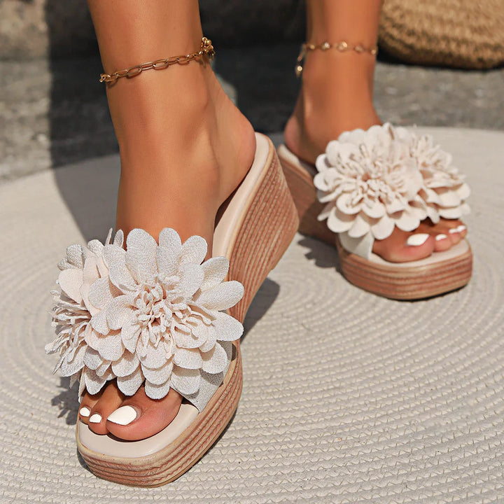 Woman wearing beige wedge sandals with large flower decorations on toes and gold anklets