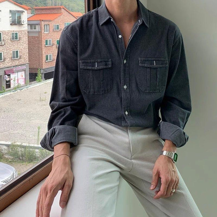 Man wearing dark denim shirt with chest pockets and light beige pants sitting by window
