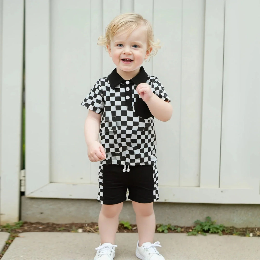 Smiling toddler boy in black and white checkered shirt and shorts standing outdoors near a white fence