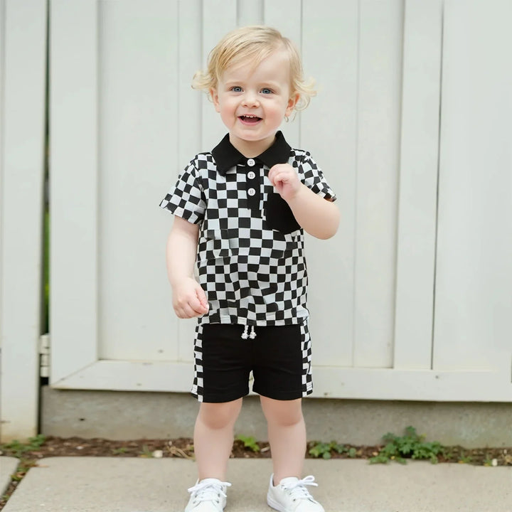 Smiling toddler boy in black and white checkered shirt and shorts standing outdoors near a white fence