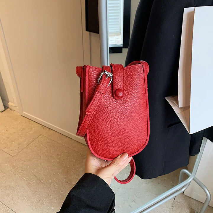 Red textured leather crossbody bag with buckle strap and snap button, held in hand indoors