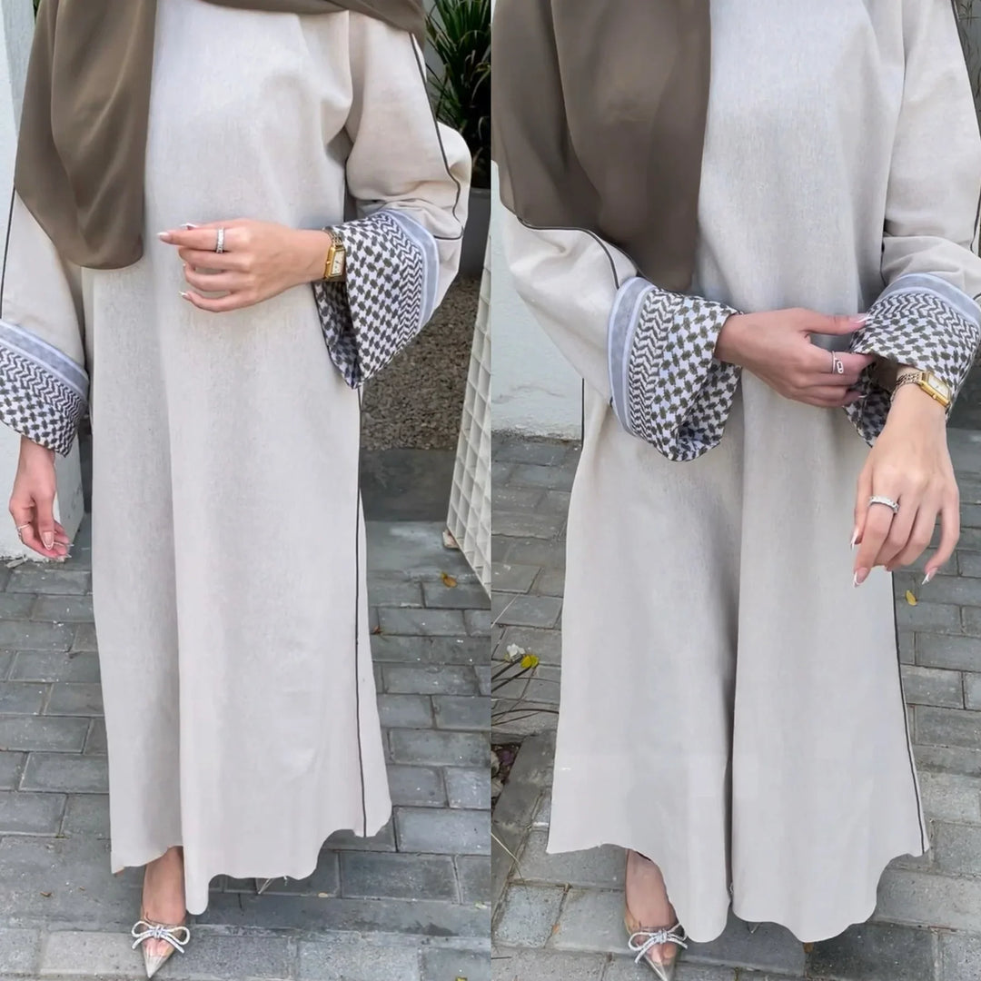 Woman wearing elegant beige abaya with patterned cuffs and taupe hijab, standing on outdoor paved path