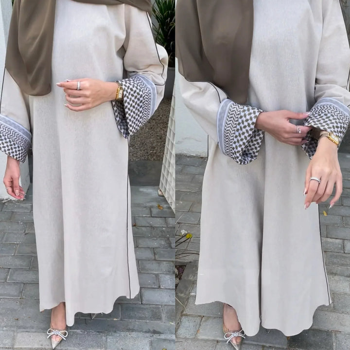 Woman wearing elegant beige abaya with patterned cuffs and taupe hijab, standing on outdoor paved path