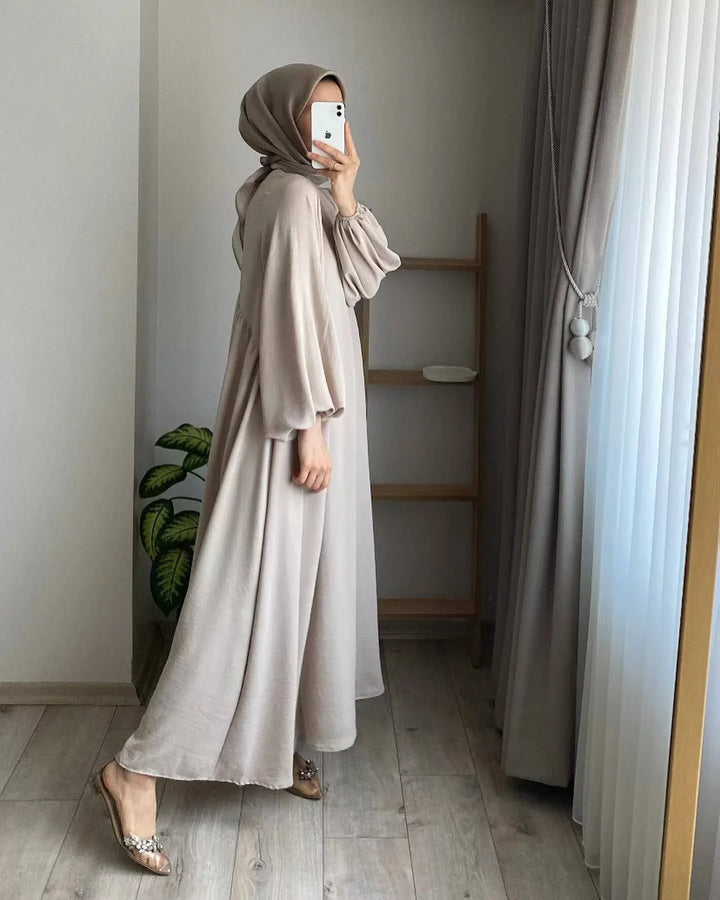 Woman wearing beige abaya and hijab taking mirror selfie in minimalistic room with wooden floor