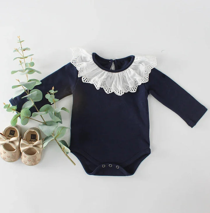 Navy blue baby onesie with white lace collar and gold baby shoes with bows on white background