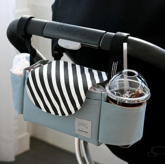 Blue stroller organizer attached to black stroller handle holding a black and white striped cloth, baby bottle, and a plastic cup with iced drink