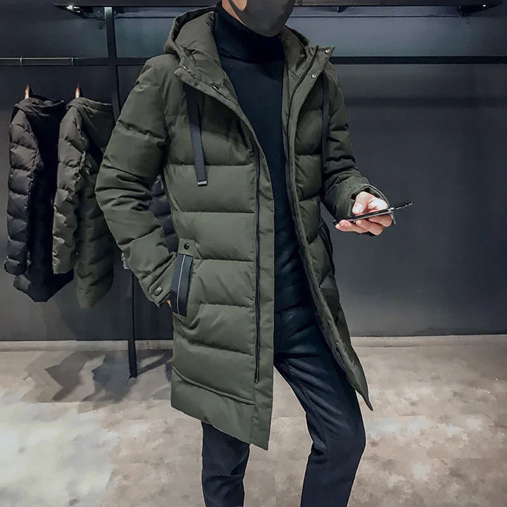 Man wearing olive green long quilted winter coat with hood, black turtleneck and pants, holding phone indoors