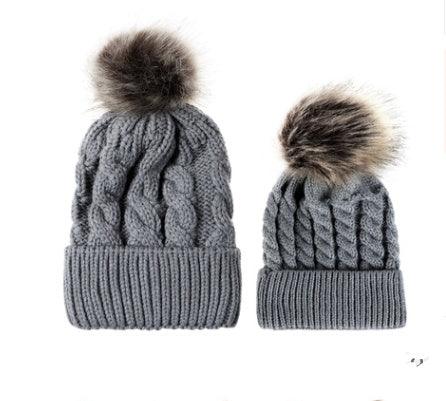 Pair of gray knit cable beanie hats with faux fur pom-poms, adult and child sizes