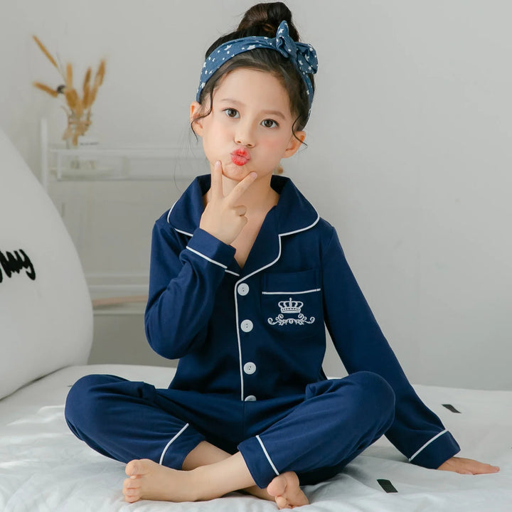 Little girl sitting cross-legged on bed wearing navy blue pajamas with white piping and crown embroidery